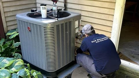 E&A technician kneeling at a Rheem condensing unit tucked between hostas and yellow siding, gauges set on top of the unit