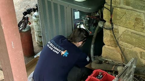 E&A technician crouched beside an older outdoor condenser at a stone wall with a red tool case open on the ground